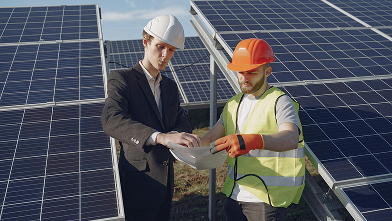 Two workers wearing helmets and discussing a tablet in front of solar panels.