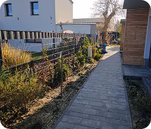 Gepflasterter Gehweg entlang eines Hauses mit einem Drahtzaun und Pflanzen auf der linken Seite.