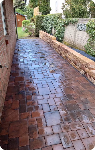 Nasse gepflasterte Terrasse neben einem Ziegelhaus mit einer niedrigen Steinmauer und grünen Hecken.