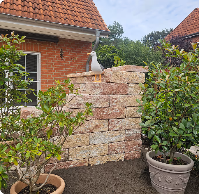 Eine Terrasse mit Mauer aus beigem Ziegelstein, zwei Töpfe mit grünen Pflanzen und eine Plastikmöwe auf der Mauer.