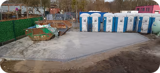 Baustelle mit neu verlegten Betonplatten, Müllcontainern und einer Reihe von blauen mobilen Toiletten im Hintergrund.