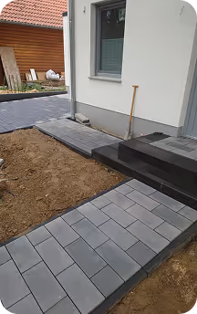 Gehweg aus grauen Betonplatten neben einem weißen Haus, daneben eine Fläche mit Erde und eine Gartenschaufel lehnt an der Hauswand.