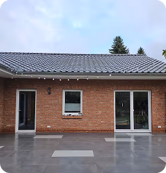 Fassade eines modernen Hauses mit rotem Ziegelmauerwerk, zwei Fenstern und einer Glastür, davor grauer Steinboden.