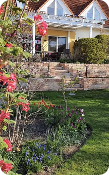 Haus mit gelber Fassade, Terrasse und gepflegtem Garten mit blühenden Blumen und grünem Rasen.