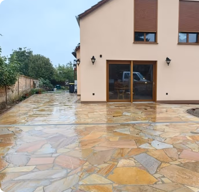 Haus mit beigem Außenputz, großen Fenstern mit braunen Rollläden und nassem, mehrfarbigem Steinboden im Hof an einem bewölkten Tag.