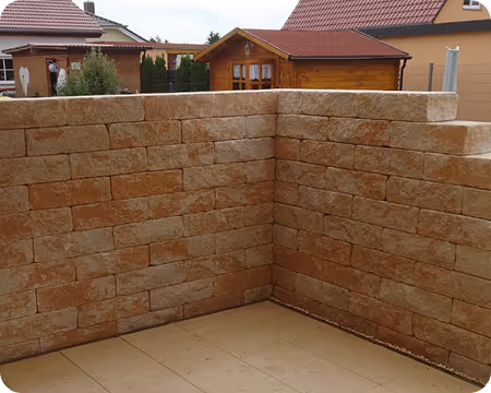 Ecke eines Gartens mit einer Winkelmauer aus gelben Natursteinen und Fliesenboden im Vordergrund.