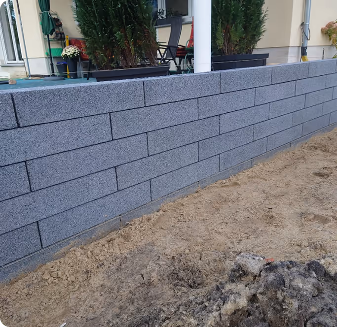 Graue Steinmauer aus rechteckigen Blöcken vor einem Haus mit Terrasse und Pflanzen.