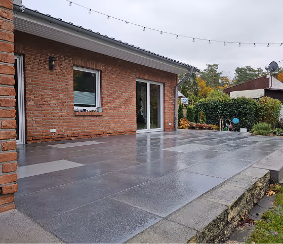 Große Terrasse mit grauen Fliesen vor einem Backsteinhaus unter einem bewölkten Himmel.