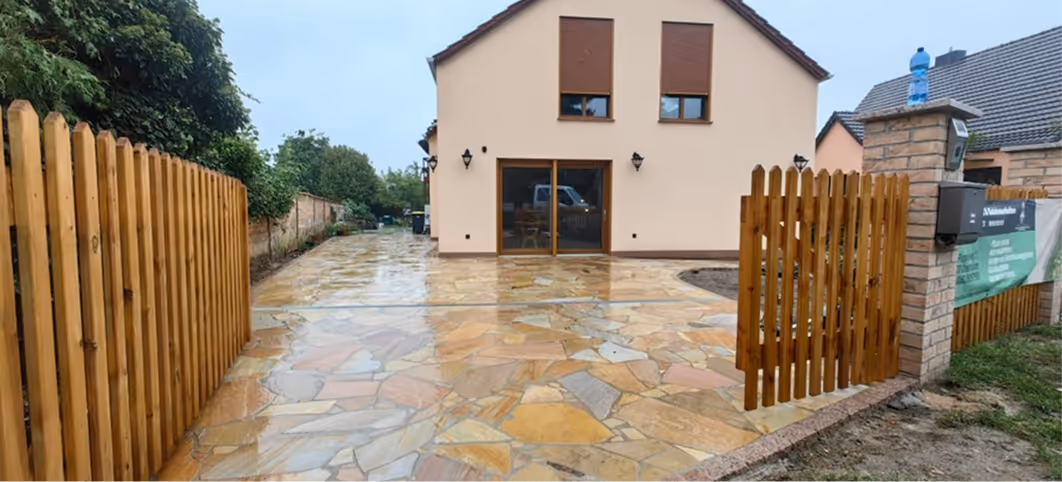 Haus mit cremefarbener Fassade, zwei Fenstern und einer Glastür, davor ein mit Naturstein belegter, nasser Gehweg und ein offenes Holztor.