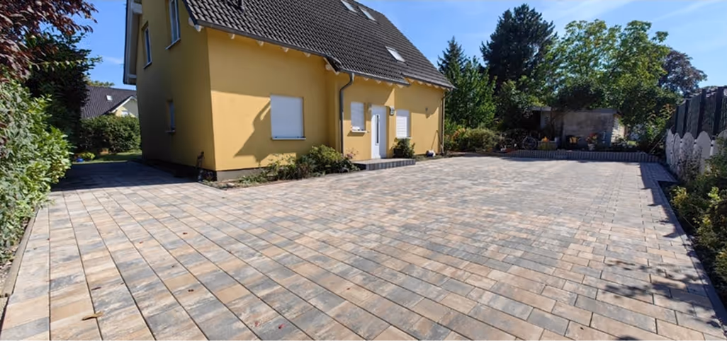 Große gepflasterte Einfahrt neben einem zweistöckigen gelben Haus mit weißen Fensterläden, umgeben von Büschen und Bäumen.