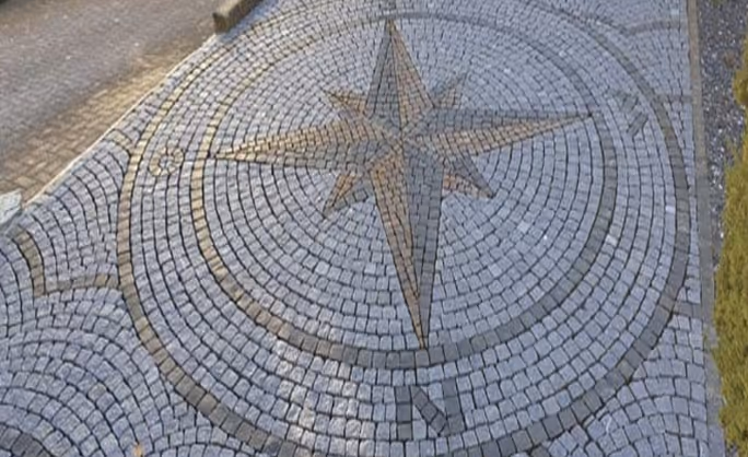 Kreisförmiges Pflastermuster mit einer Kompassrose aus grauen Steinplatten auf einem Bürgersteig.