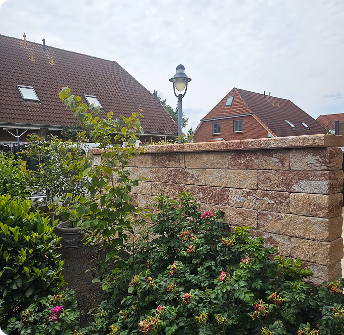 Garten mit blühenden Sträuchern, einer steinernen Mauer, Häusern mit roten Ziegeldächern und einer Straßenlaterne im Hintergrund.