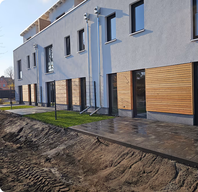 Moderne zweistöckige Wohnhäuser mit weißen Wänden, Holzverkleidungen und Terrassen neben einem frisch ausgehobenen Erdgraben.