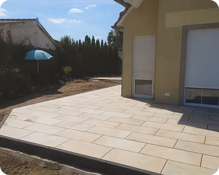 Sonnige Terrasse mit hellen rechteckigen Fliesen neben einem Haus mit geschlossenen Rollläden.