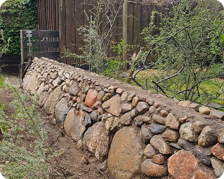 Handgebaute Natursteinmauer mit unterschiedlicher Größe und Form, umgeben von Garten und Büschen.