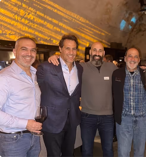 Four men smiling and standing closely together at an indoor event, one holding a glass of red wine.