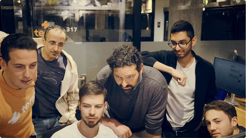 Group of six men gathered around a computer screen in a modern office setting, focused on the display.