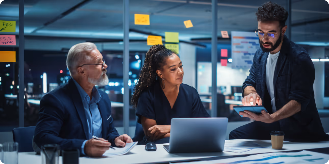 Three colleagues collaborating at a table in a modern office at night, with one person showing information on a tablet while others listen attentively.