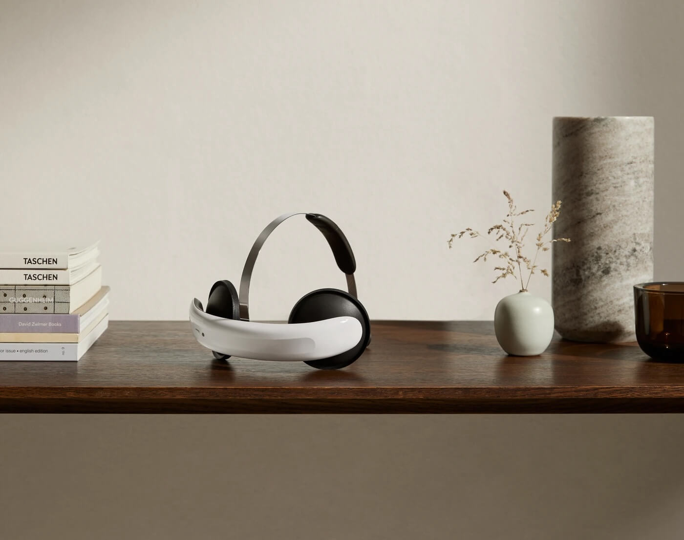 Flow tDCS-headset, ett vitt band med svarta öronkåpor och en tunn metallbygel, står på en träbänk bredvid en bokstapel, en liten vas med torkade strån och en marmorvas.