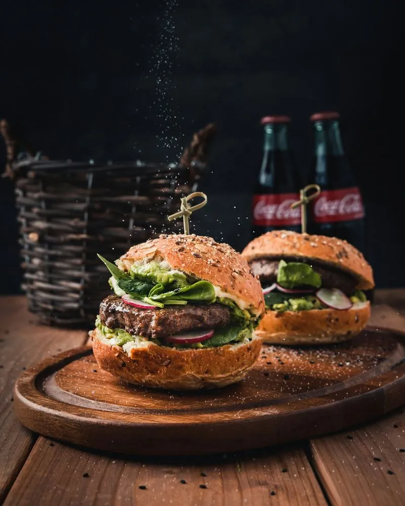 Due hamburger con pane ai semi, carne, foglie di spinaci e fette di ravanello su un tagliere di legno, con due bottiglie di Coca-Cola sullo sfondo.