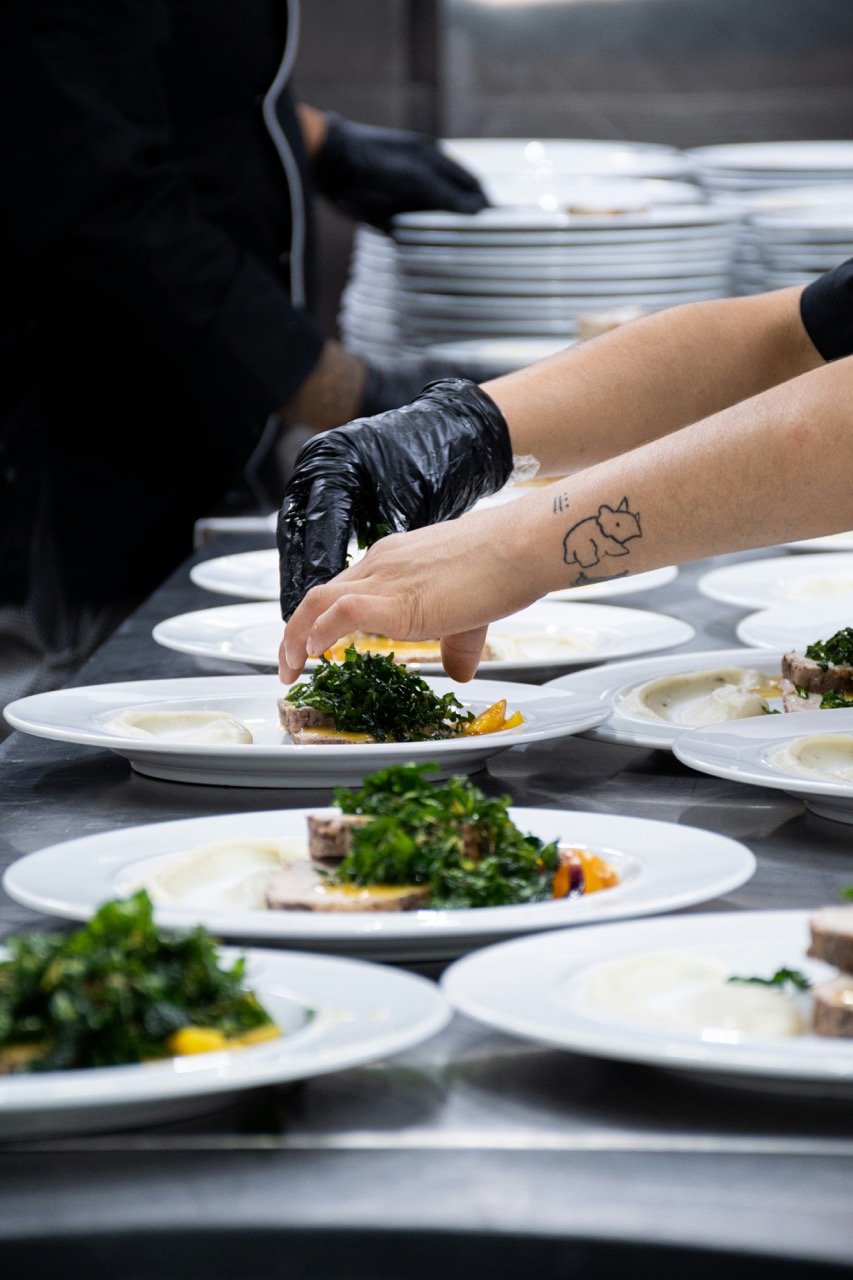 Chef wearing black gloves garnishing plates with leafy greens and meat in a professional kitchen.