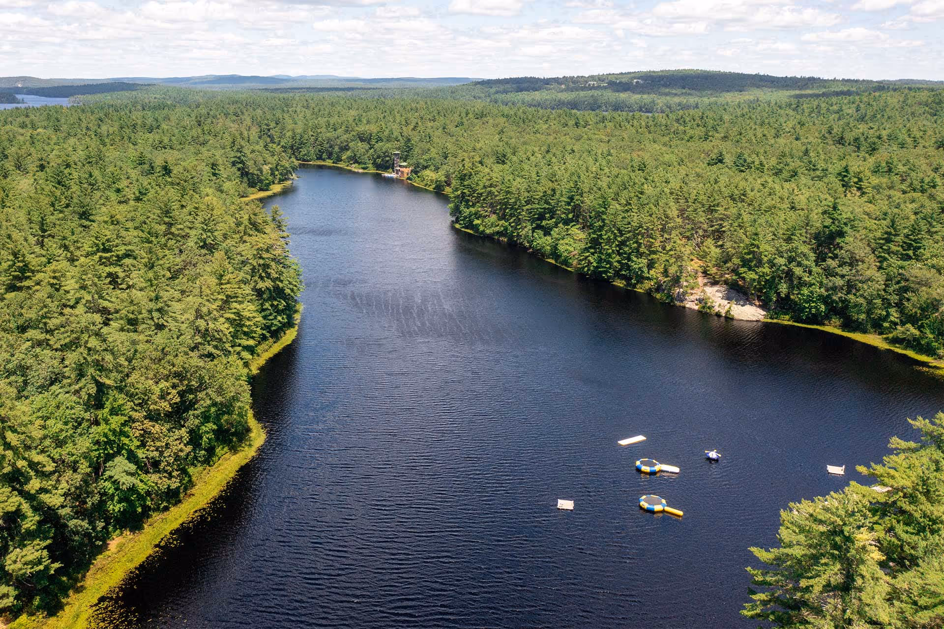 Aerial view of a narrow lake surrounded by dense forest with several floating docks and boats on the water.