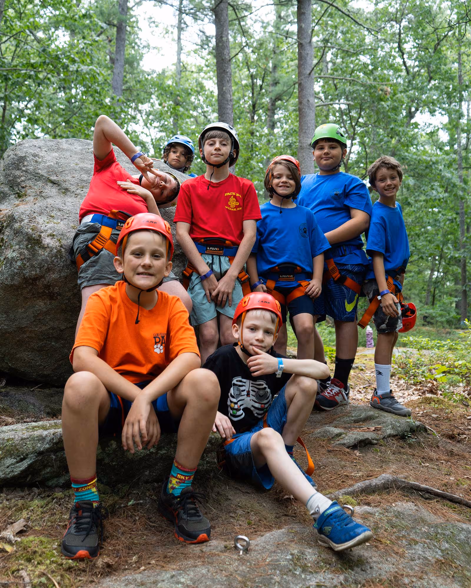 A group of eight kids in helmets and harnesses pose together in a forested area near large rocks.