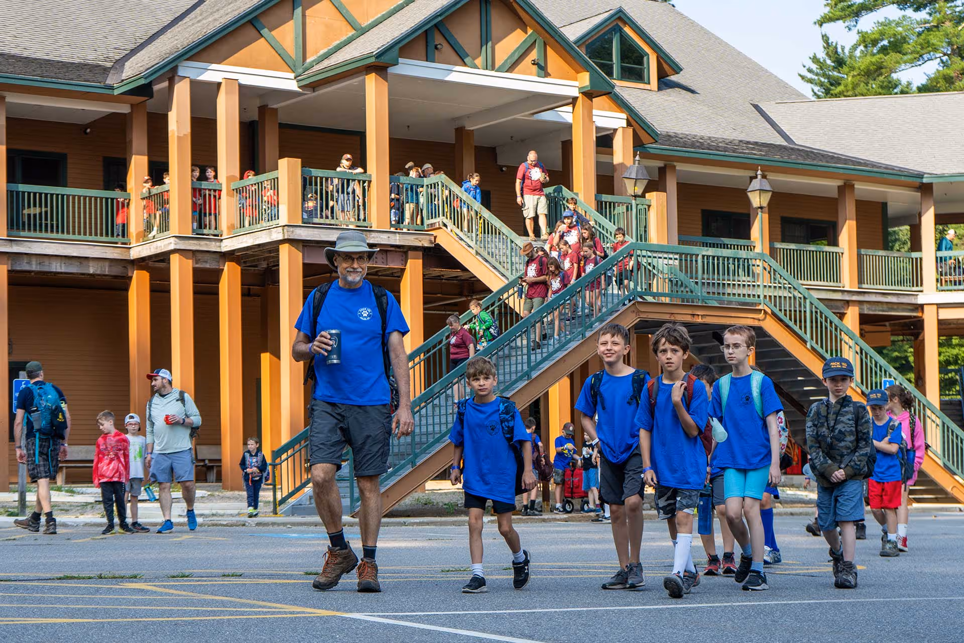 A group of boys in blue shirts walks with an adult in front of a lodge, with more people visible on the stairs and balcony.