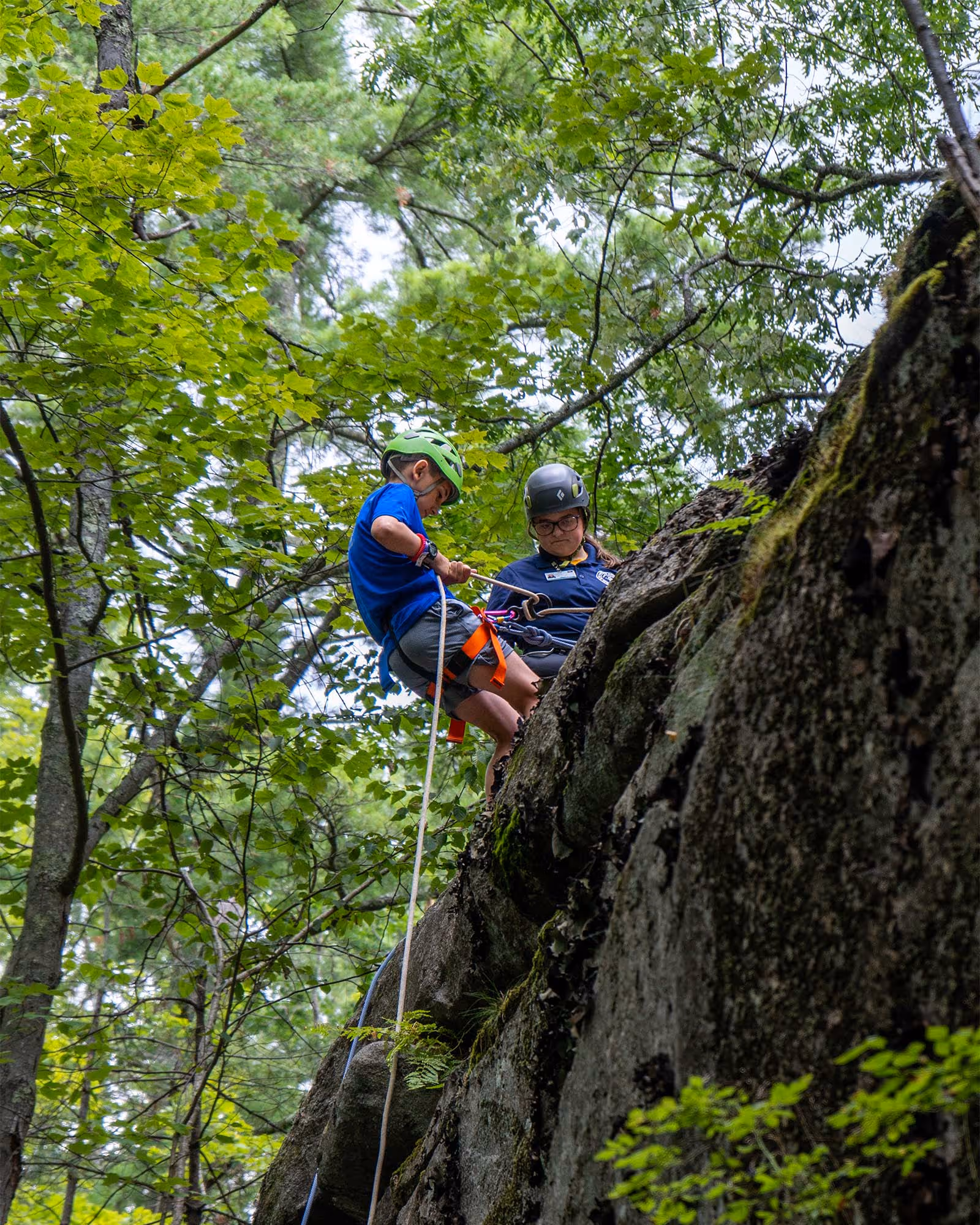 Two people wearing helmets rappel down a rocky cliff surrounded by green trees in an outdoor setting.