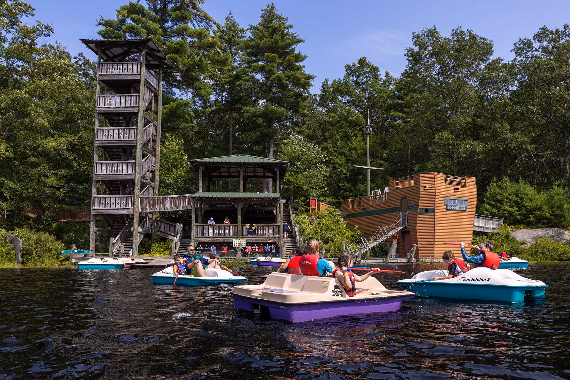 People in paddle boats on a lake near a wooden tower and a play structure shaped like a ship, surrounded by trees.