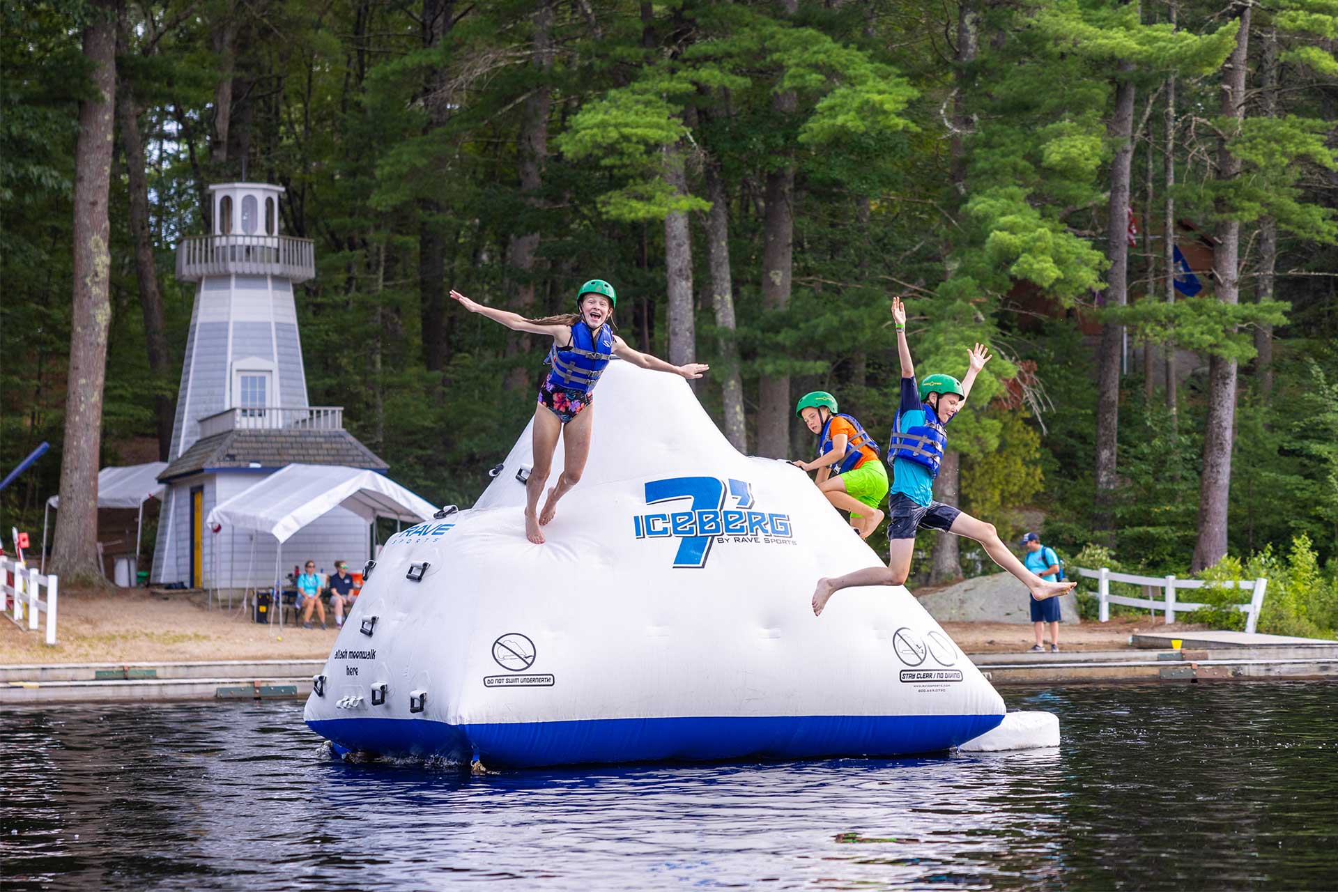 Three children wearing life jackets and helmets jump off a floating inflatable iceberg on a lake near a wooded shore.