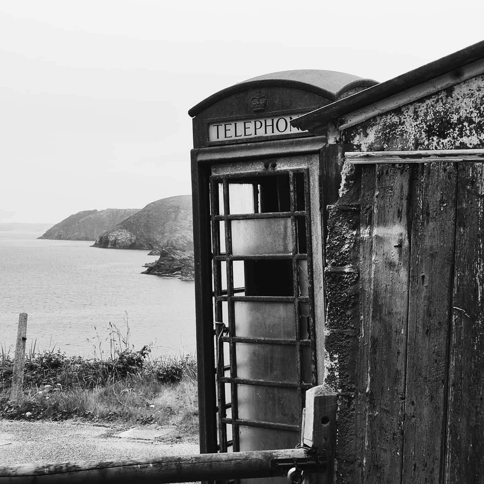 Black and white photograph of a derelict telephone box with broken panes standing on a clifftop above the sea.
