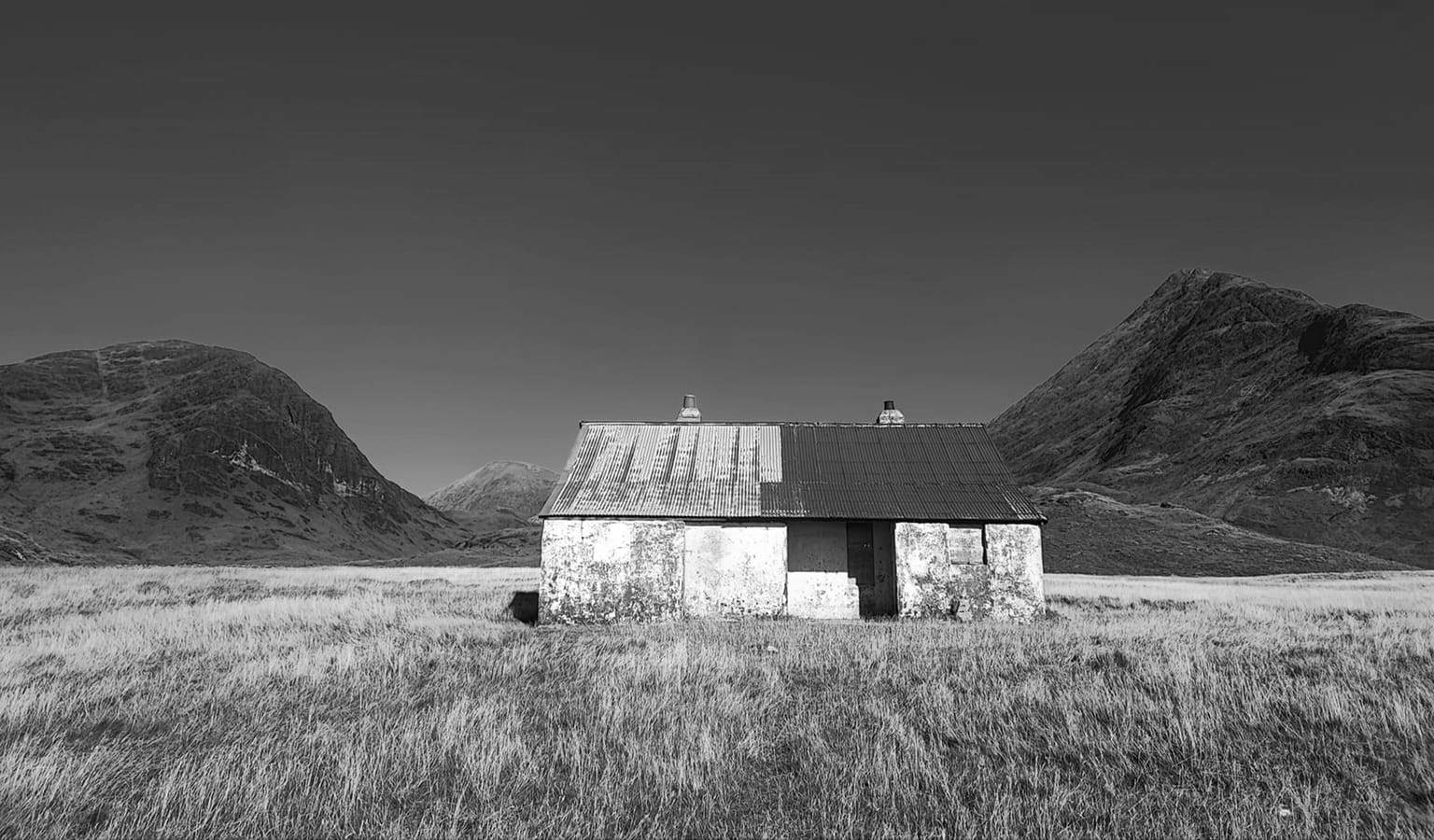 Black and white photograph of a small stone cottage with a corrugated roof standing alone in a wide moorland valley between hills.