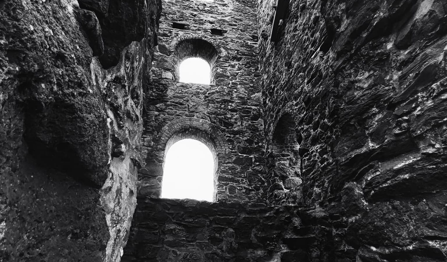 Black and white photograph looking up inside a ruined stone tower, two arched windows letting in bright light.