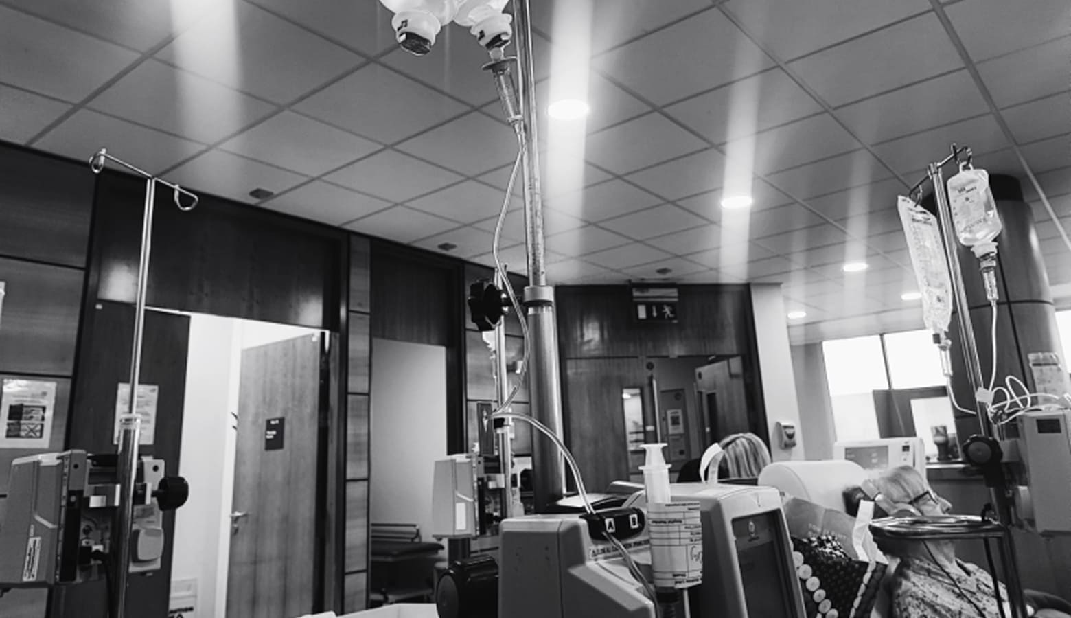 Black and white photograph of a hospital ward with IV drip stands and monitors beside empty treatment chairs.