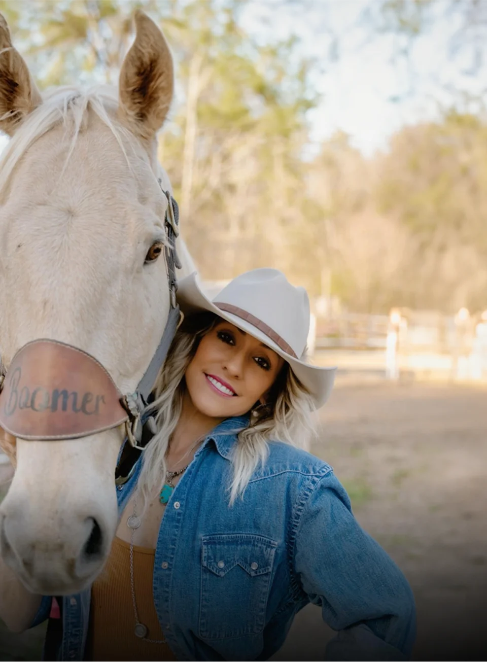 Smiling woman in a white cowboy hat and denim jacket standing next to a white horse wearing a leather noseband engraved with 'Boomer'.