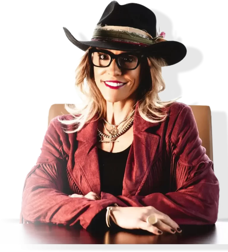 Smiling woman wearing a black hat, glasses, and a red jacket, sitting at a table with arms crossed.