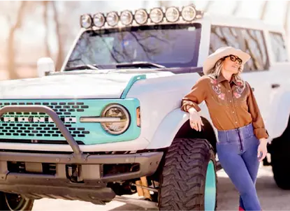 Woman in a hat and sunglasses leaning against a white and turquoise off-road vehicle with large tires.