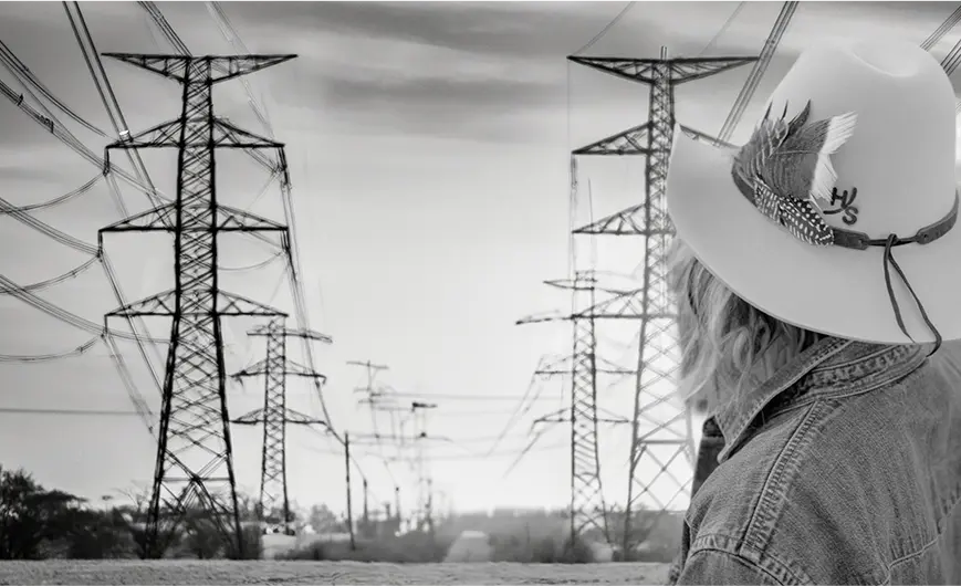 Person wearing a hat with feathers and a denim jacket looking at a row of electrical transmission towers in a landscape.