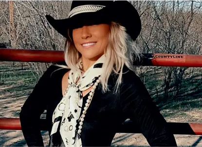 Smiling woman wearing a black cowboy hat, black shirt, and white patterned scarf standing outdoors with leafless trees and a red fence behind her.