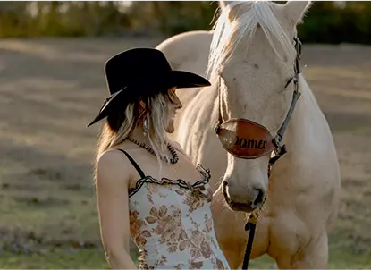 Woman wearing a black cowboy hat and floral dress standing next to a white horse in an outdoor setting.