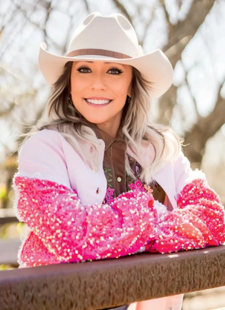 Smiling woman wearing a beige cowboy hat and a pink sparkly jacket leaning on a fence outdoors.
