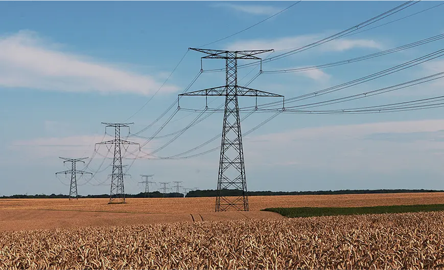 Series of electrical transmission towers standing in a golden wheat field under a blue sky with scattered clouds.