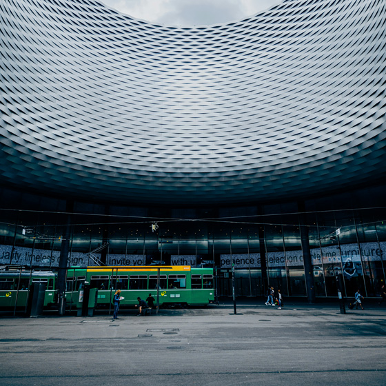 Interior of a modern circular building with a patterned ceiling and a green tram inside, with people walking nearby.