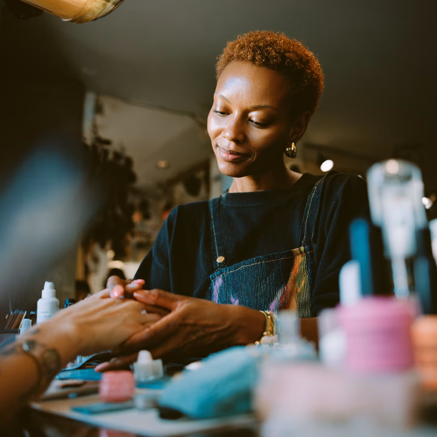 Manicurista con camiseta negra y peto azul oscuro realizando un tratamiento de uñas a una clienta en un salón de belleza.