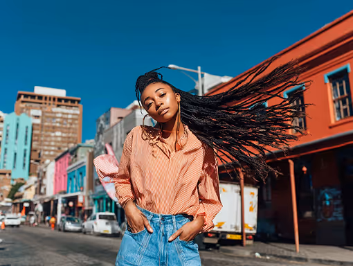 Mujer con trenzas largas que se mueve al viento, con camisa naranja y pantalones vaqueros, posando en una calle colorida de la ciudad.