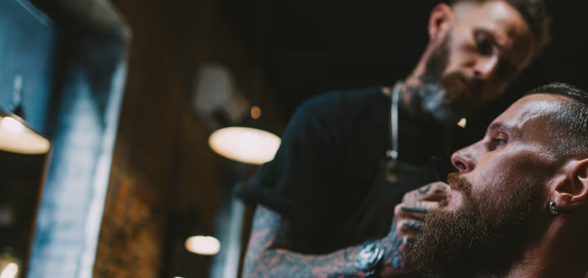 Bearded man getting a beard trim from a tattooed barber in a dimly lit barbershop.