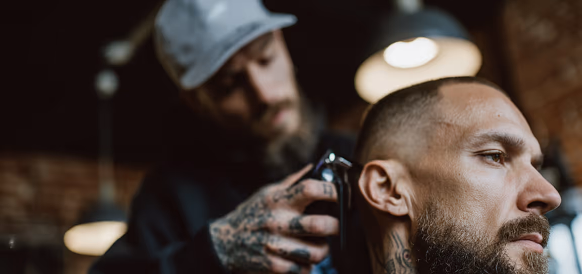 Hombre con barba y tatuajes en el cuello recibiendo un corte de cabello con máquina en una barbería.