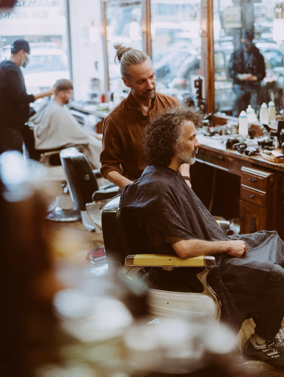 Barber cutting a middle-aged man's curly hair in a salon with another customer in the background.