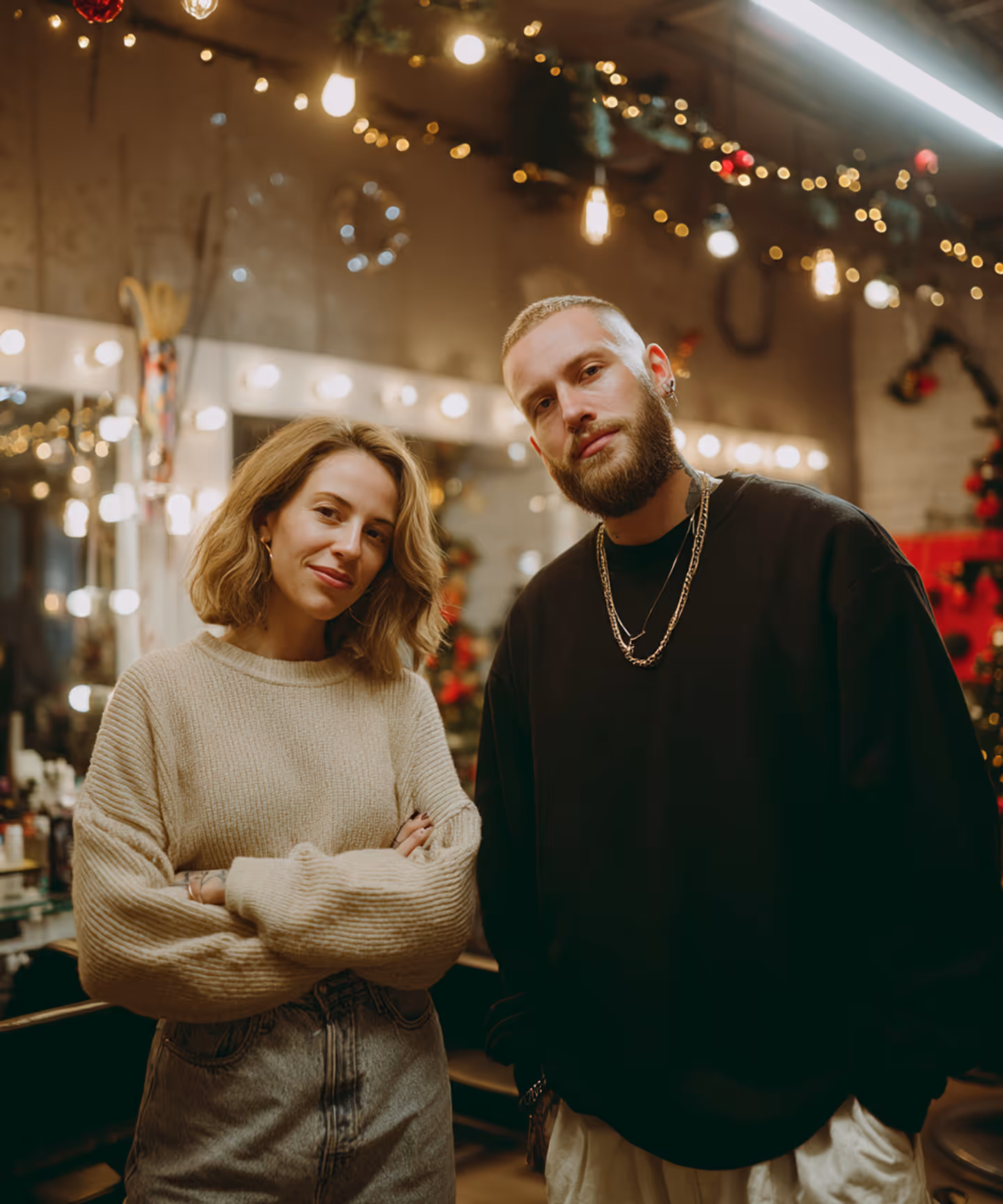 Pareja joven con suéteres casuales posando en un interior decorado con luces y adornos festivos.